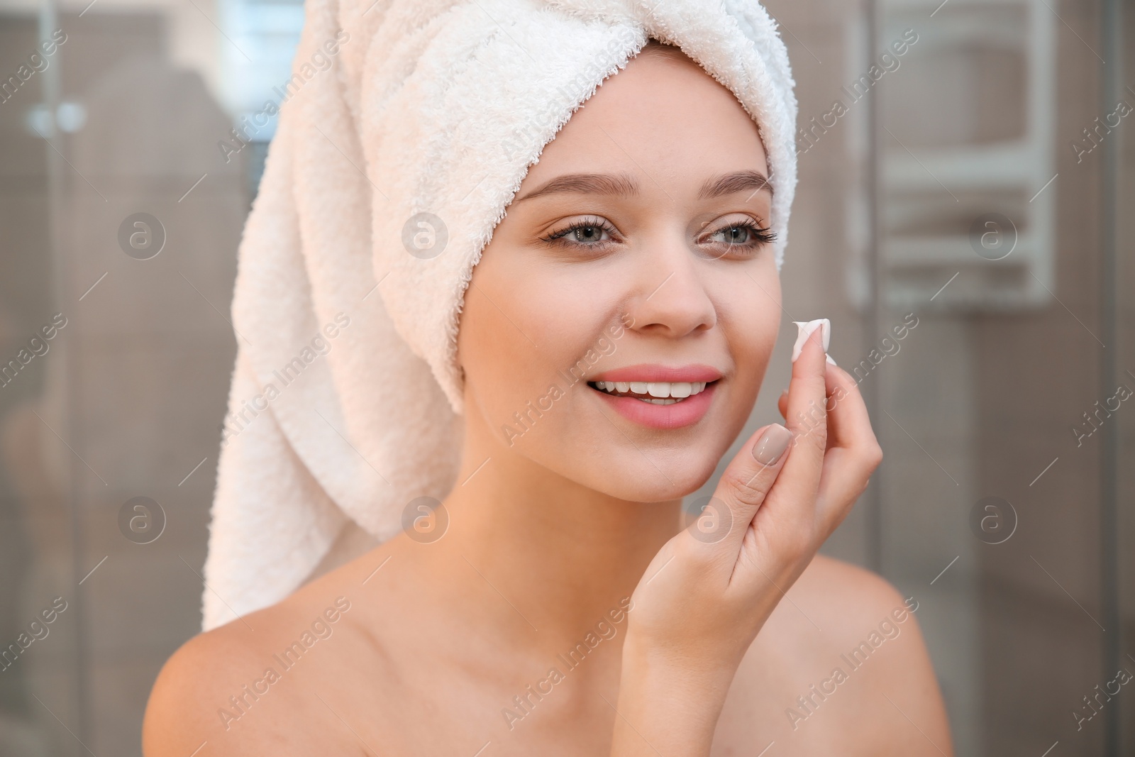 Photo of Beautiful woman with towel on head applying face cream in bathroom