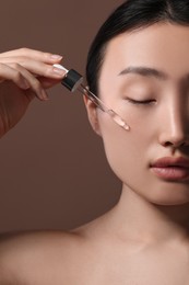 Beautiful young woman applying cosmetic serum onto her face on brown background, closeup