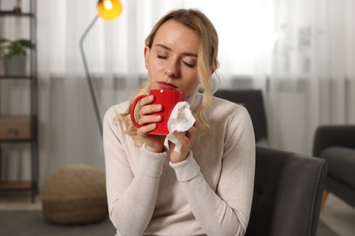Photo of Sick woman with tissue and cup of drink in armchair at home. Cold symptoms