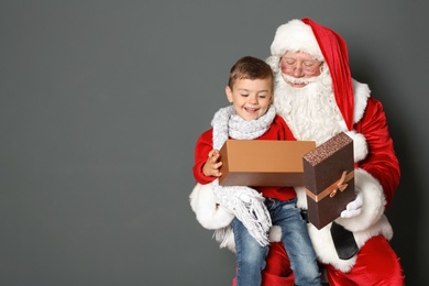 Photo of Little boy with gift box sitting on authentic Santa Claus' lap against grey background