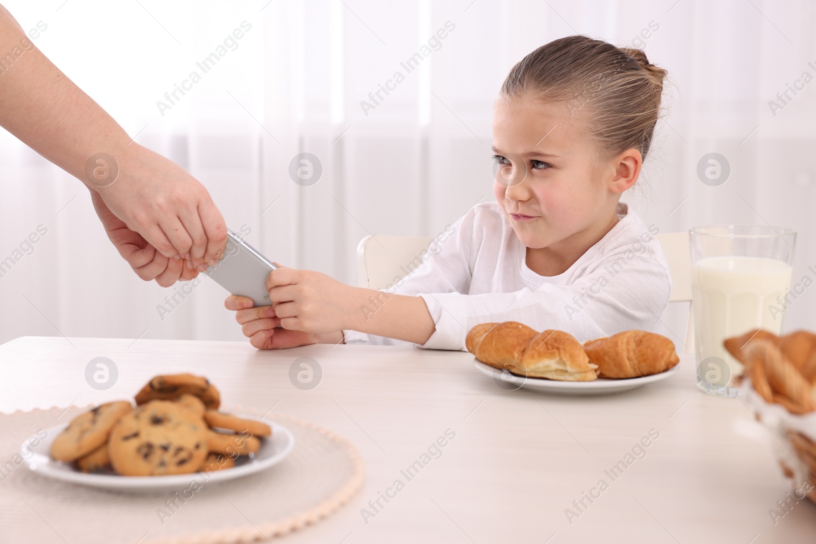 Photo of Woman taking away smartphone from her daughter at table indoors, closeup. Internet addiction