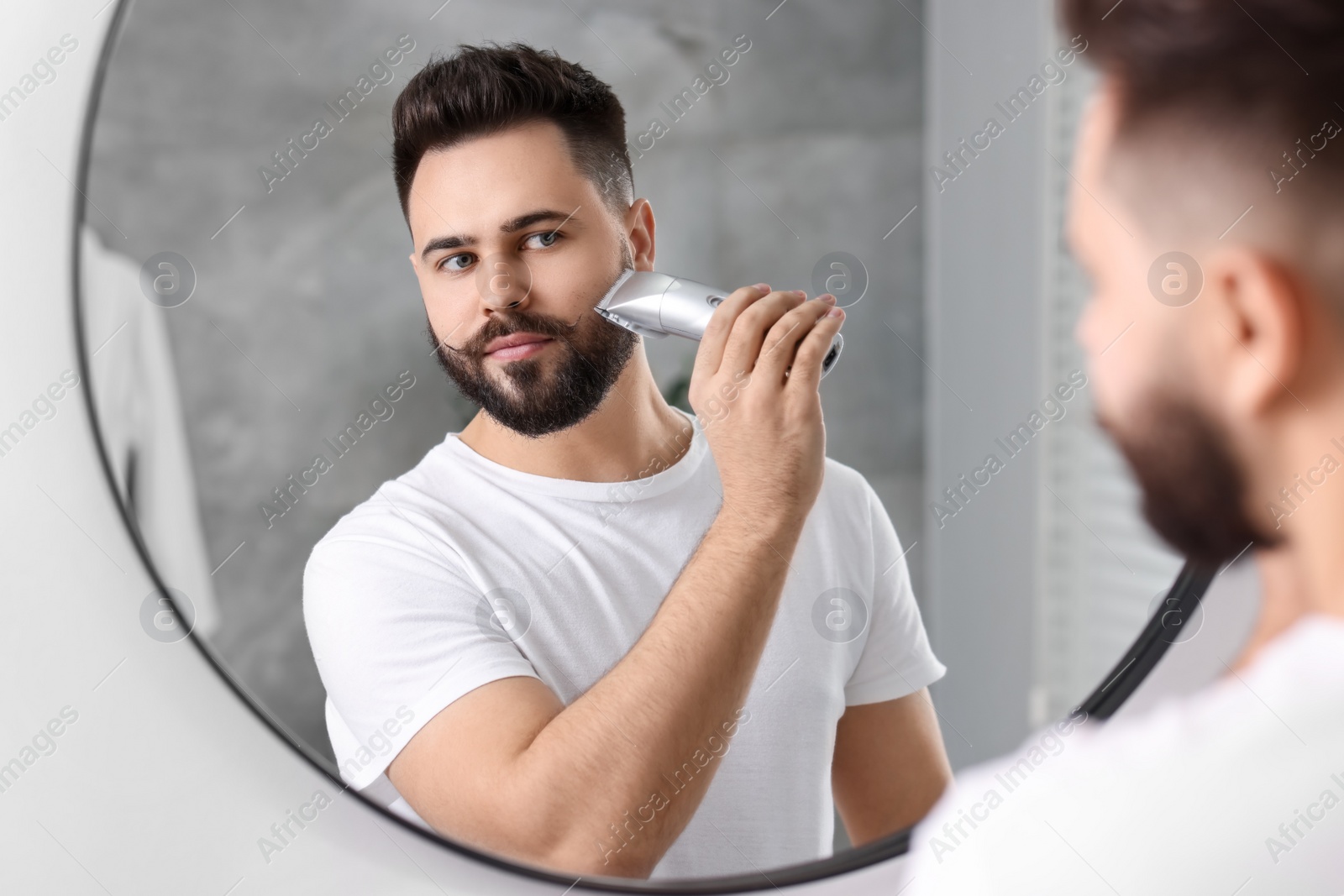 Photo of Handsome young man trimming beard near mirror in bathroom