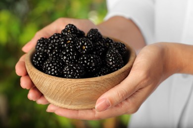 Photo of Woman holding bowl of fresh ripe black blackberries on blurred natural background, closeup