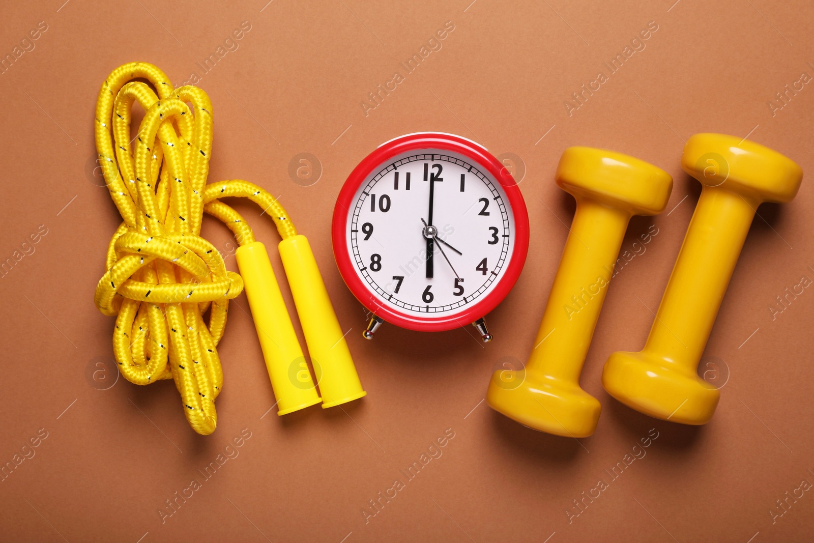Photo of Alarm clock, skipping rope and dumbbells on brown background, flat lay. Morning exercise