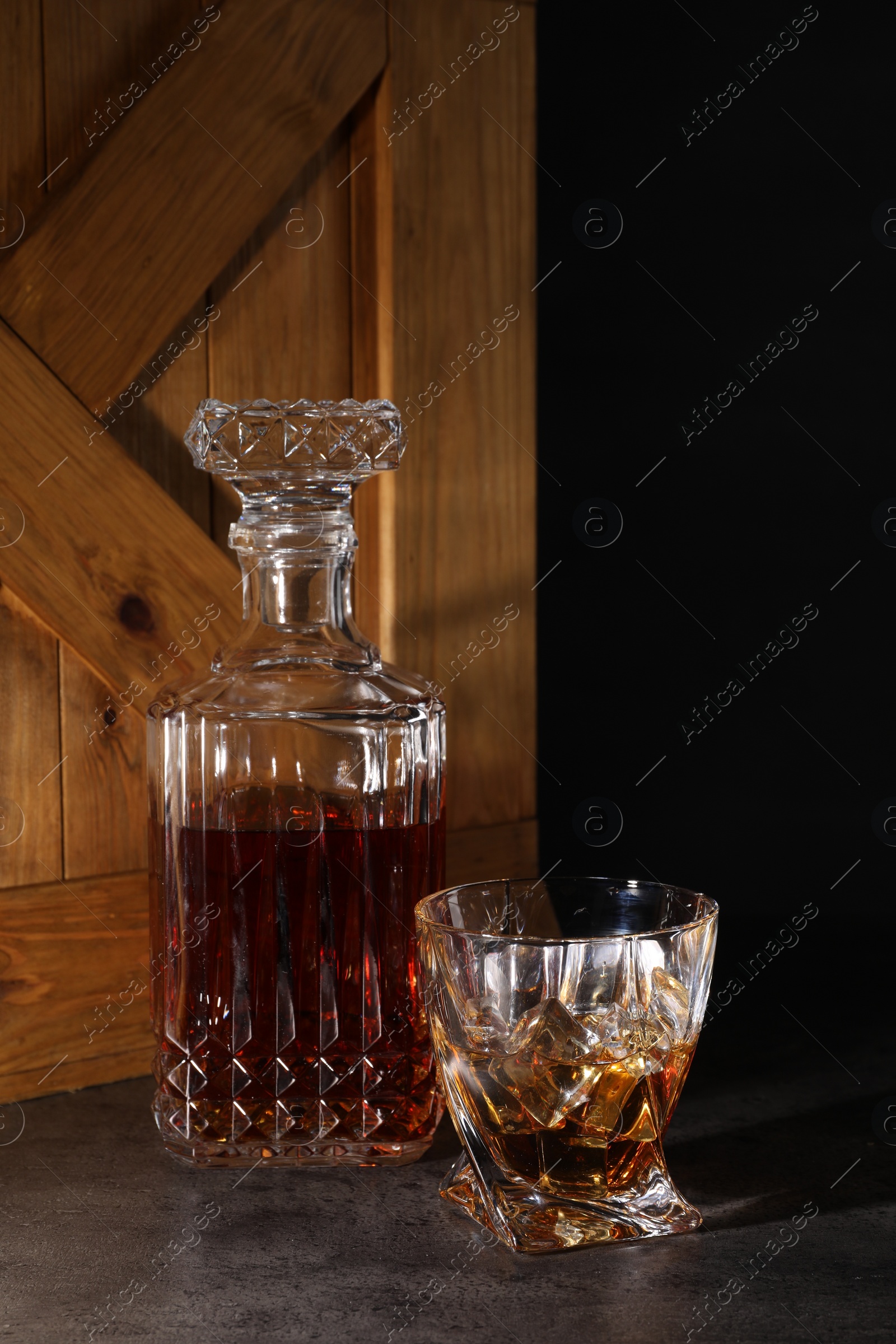 Photo of Whiskey in glass and bottle near wooden crate on dark table against black background