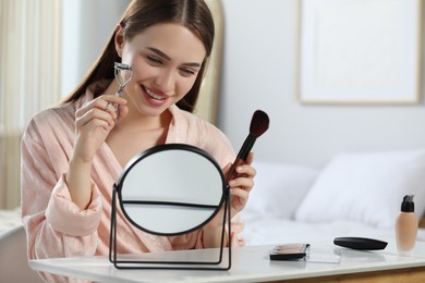 Woman with eyelash curler and brush near mirror at home