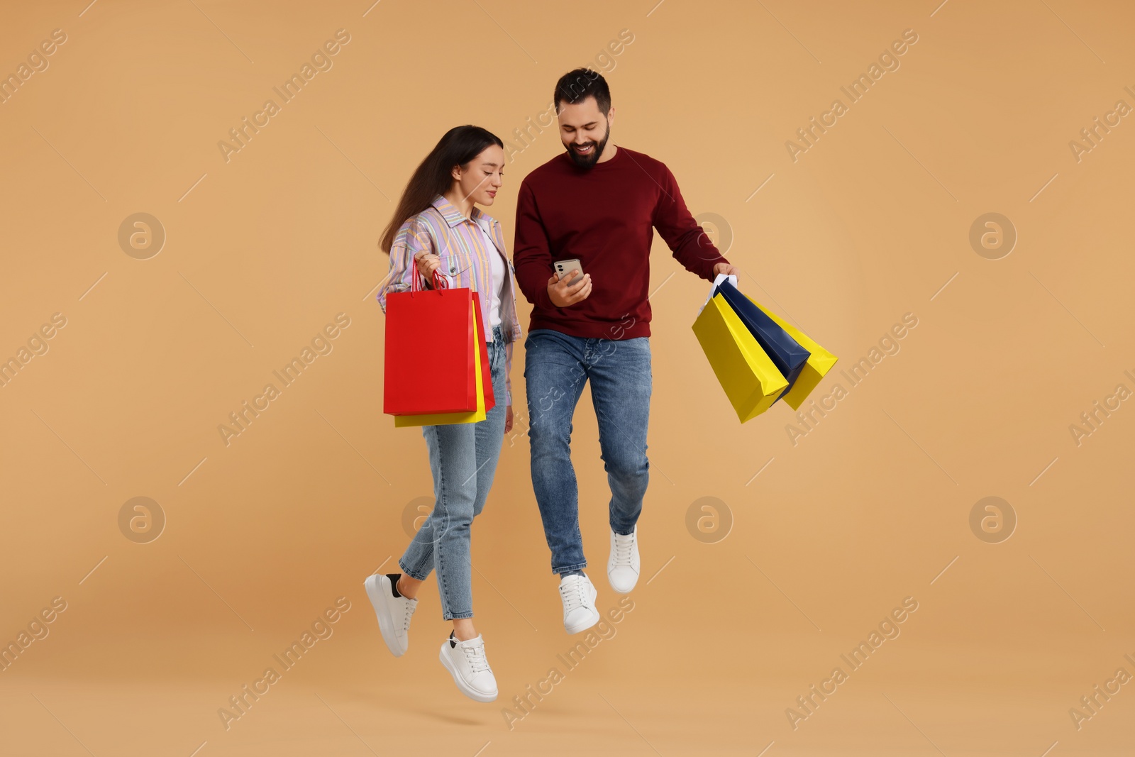 Photo of Happy couple with shopping bags and smartphone jumping on beige background