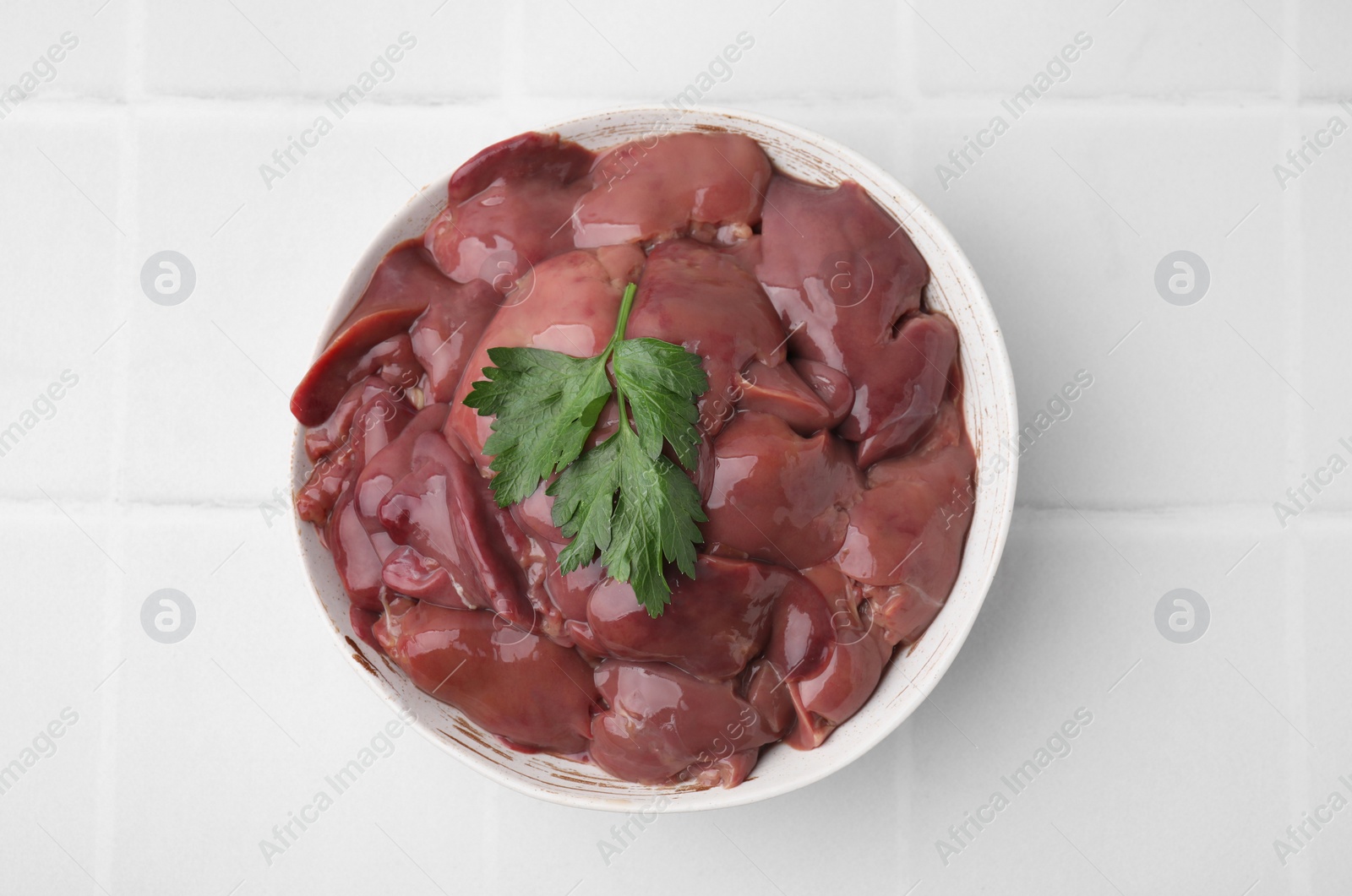 Photo of Bowl of raw chicken liver with parsley on white tiled table, top view