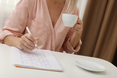 Photo of Middle aged woman with cup of drink solving sudoku puzzle at white table indoors, closeup