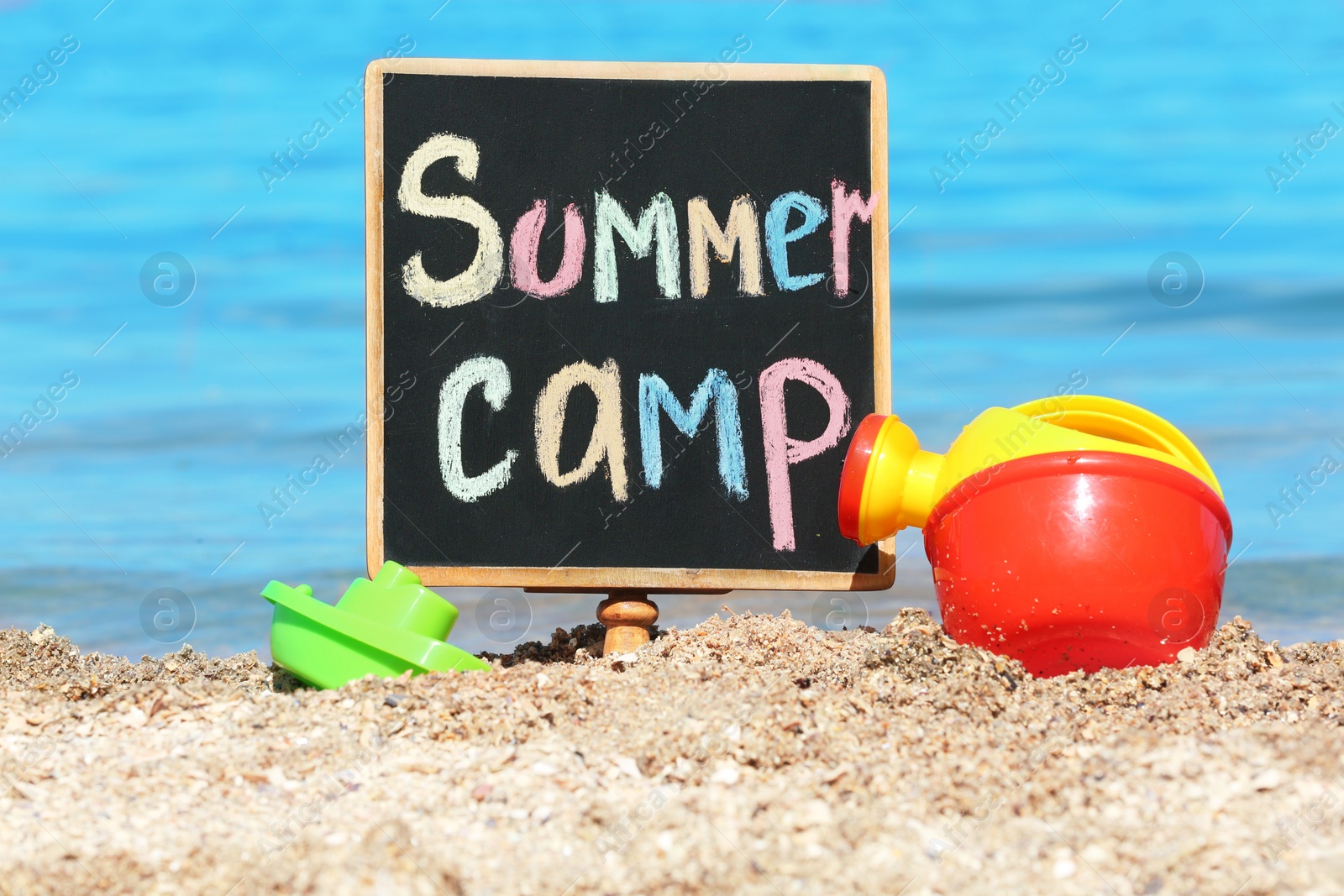 Photo of Little blackboard with text SUMMER CAMP and toys on sand near sea