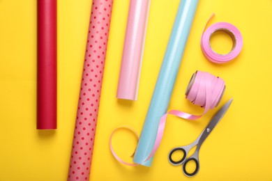 Photo of Rolls of colorful wrapping papers, scissors and ribbons on yellow background, flat lay