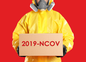 Man wearing chemical protective suit with cardboard box on red background, closeup. Coronavirus outbreak