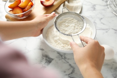 Photo of Woman sieving flour at white marble table, closeup. Cooking of delicious plum cake