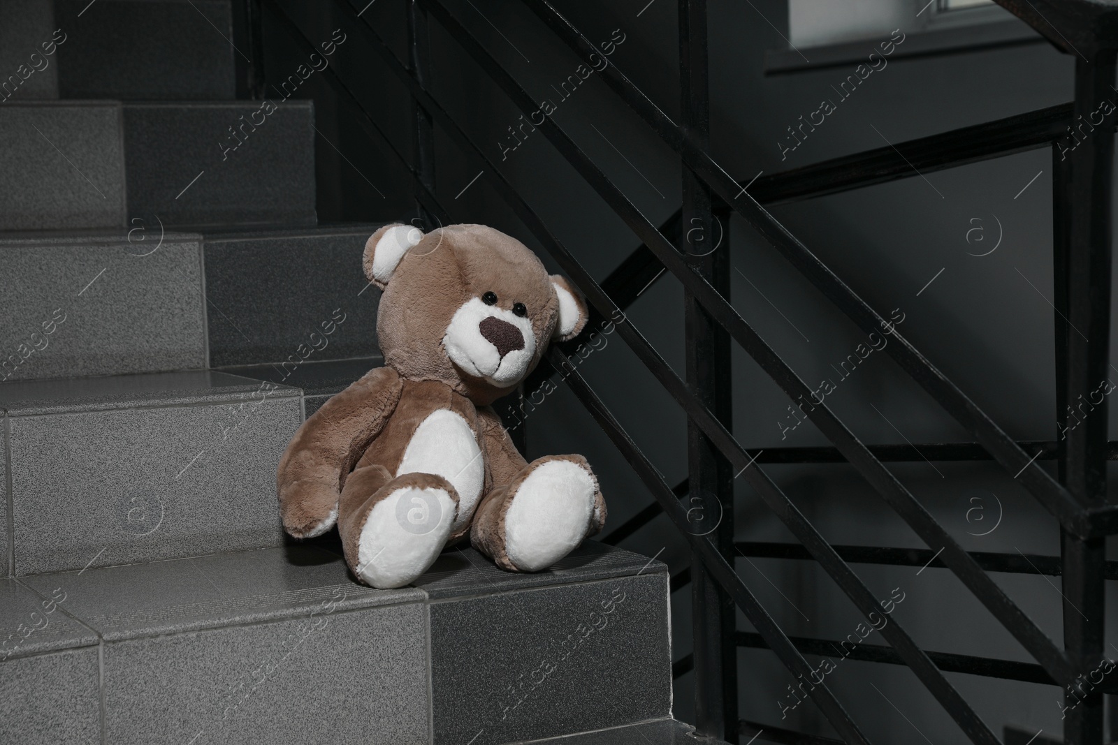 Photo of Lonely teddy bear on staircase near metal railing indoors