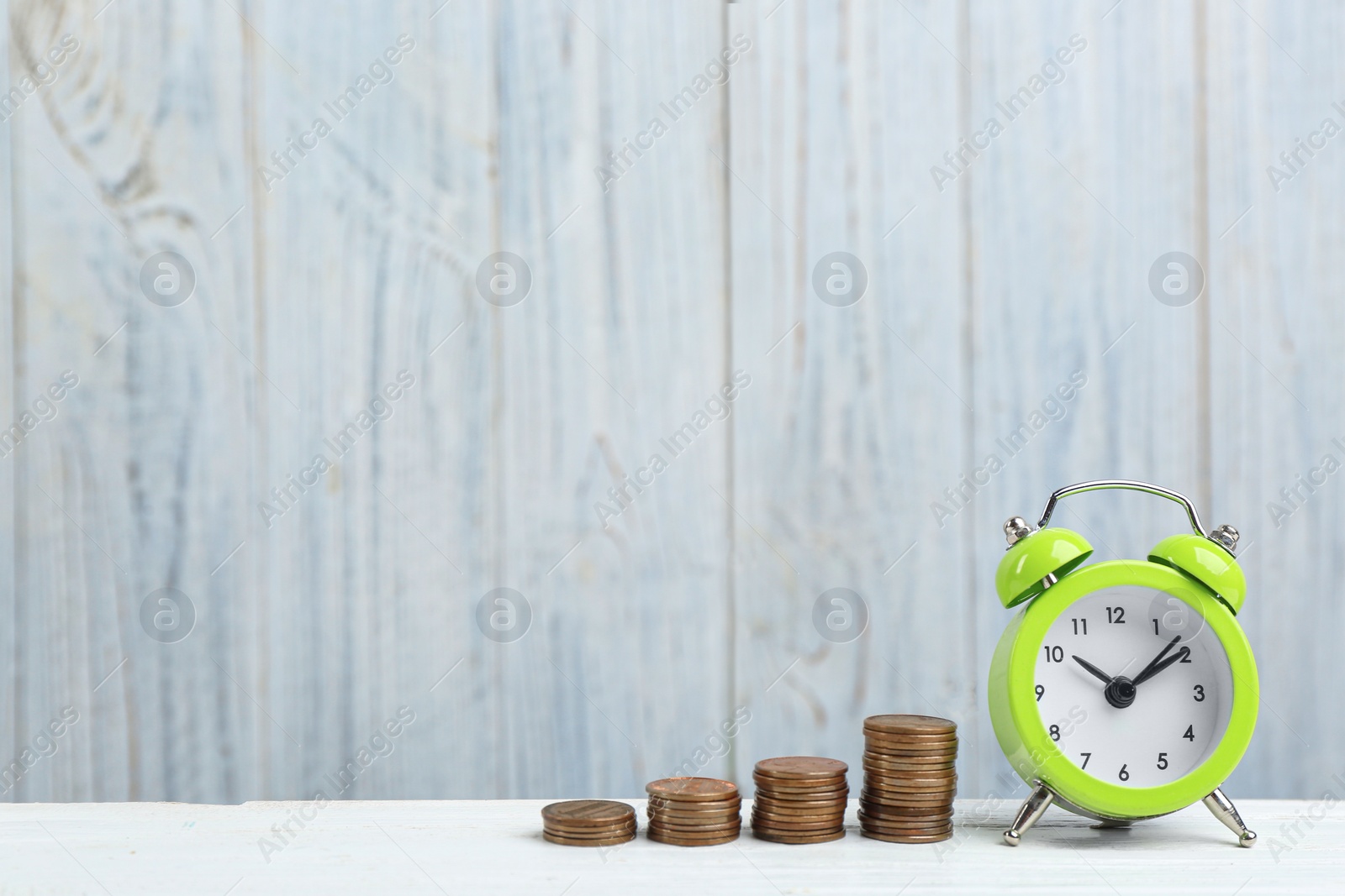 Photo of Light green alarm clock and stacked coins on white table against wooden background, space for text. Money savings