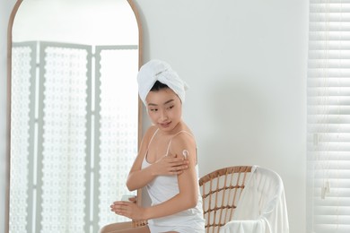 Beautiful young Asian woman applying body cream on shoulder in room