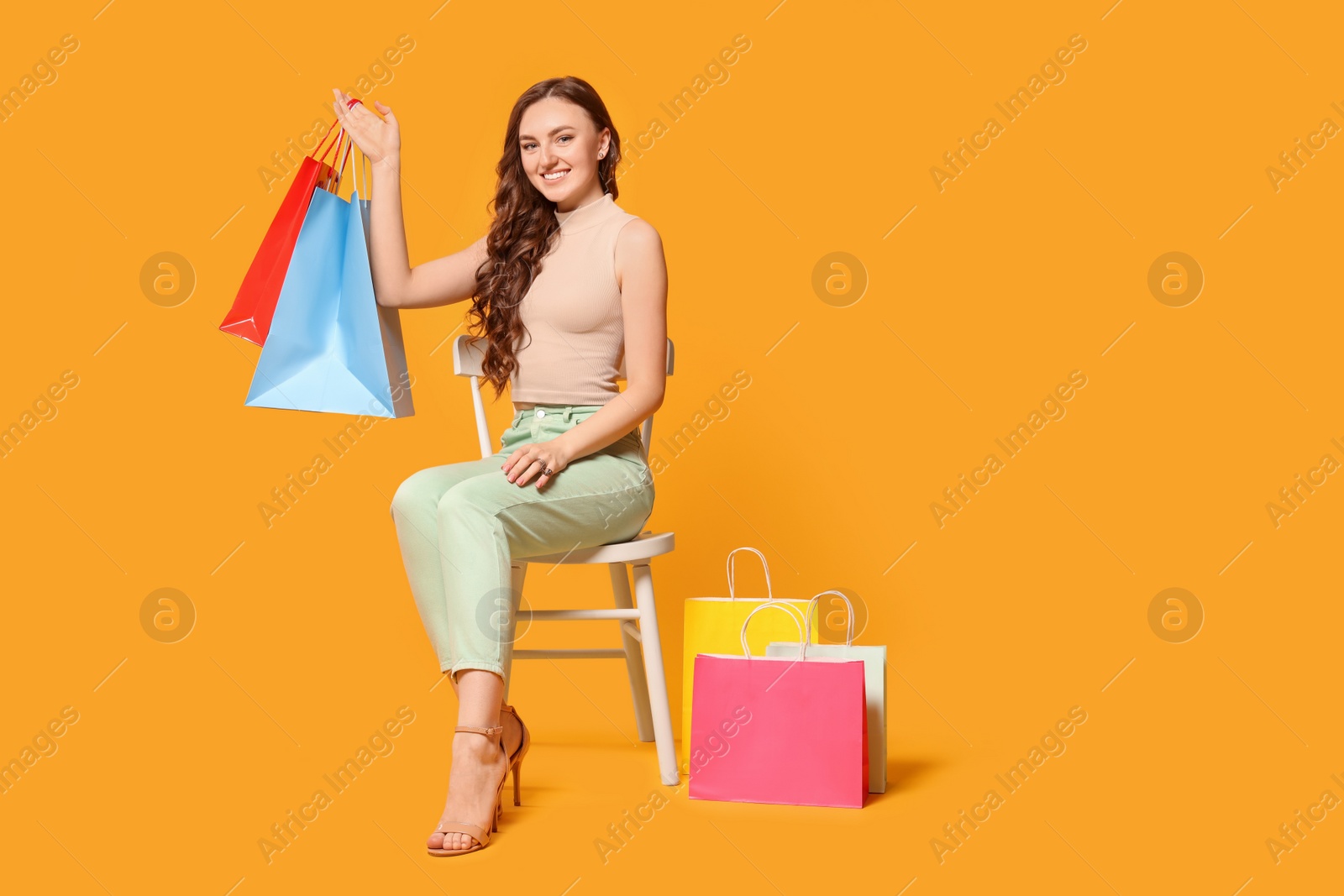 Photo of Happy woman holding colorful shopping bags on chair against orange background