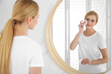 Photo of Happy young woman cleaning face with cotton pad near mirror in bathroom