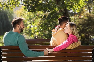 Photo of Woman holding hands with another man behind her boyfriend's back on bench in park. Love triangle