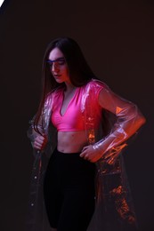Photo of Fashionable portrait of beautiful woman wearing transparent coat and glasses on dark background in neon lights