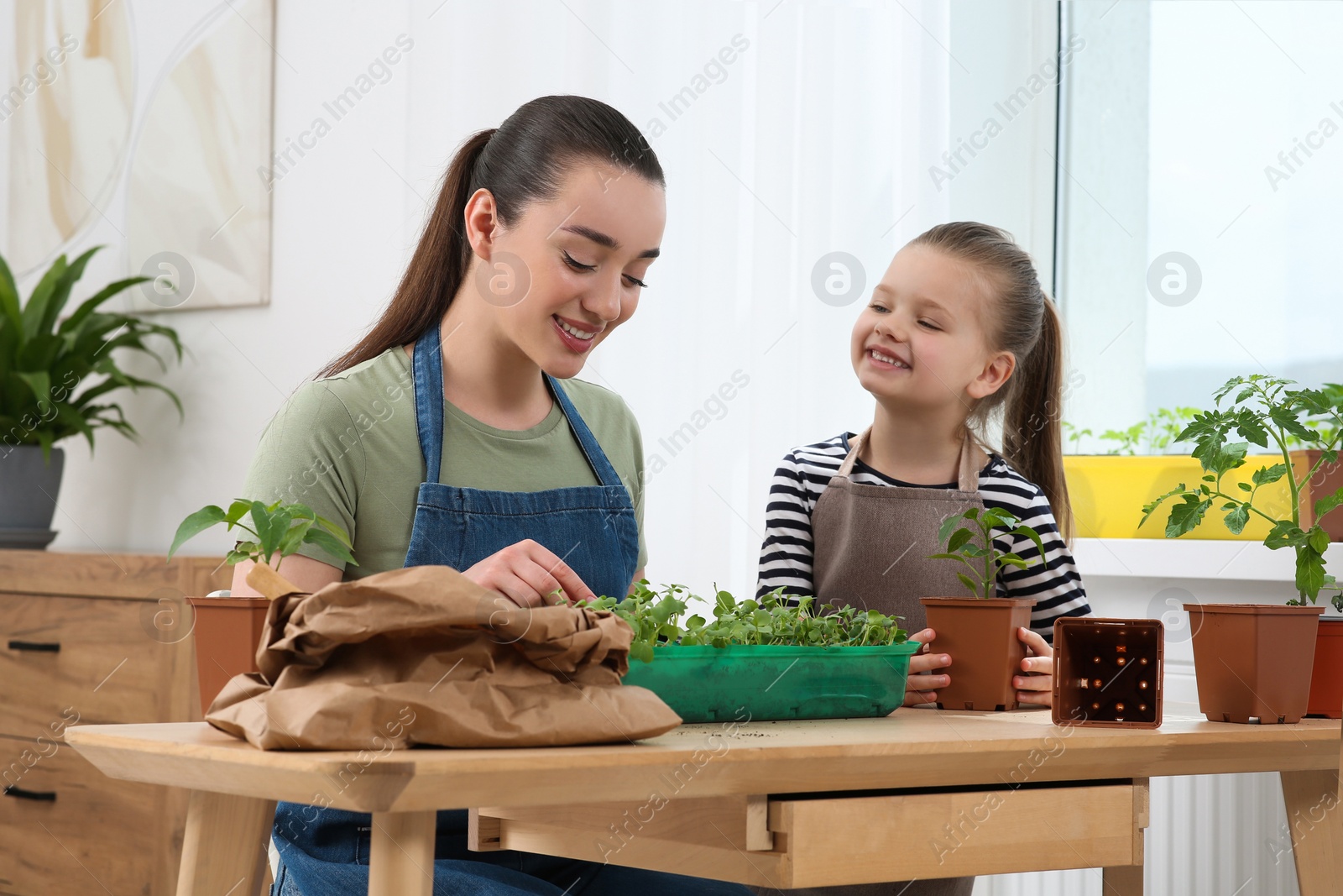 Photo of Mother and daughter planting seedlings into plastic container together at wooden table in room