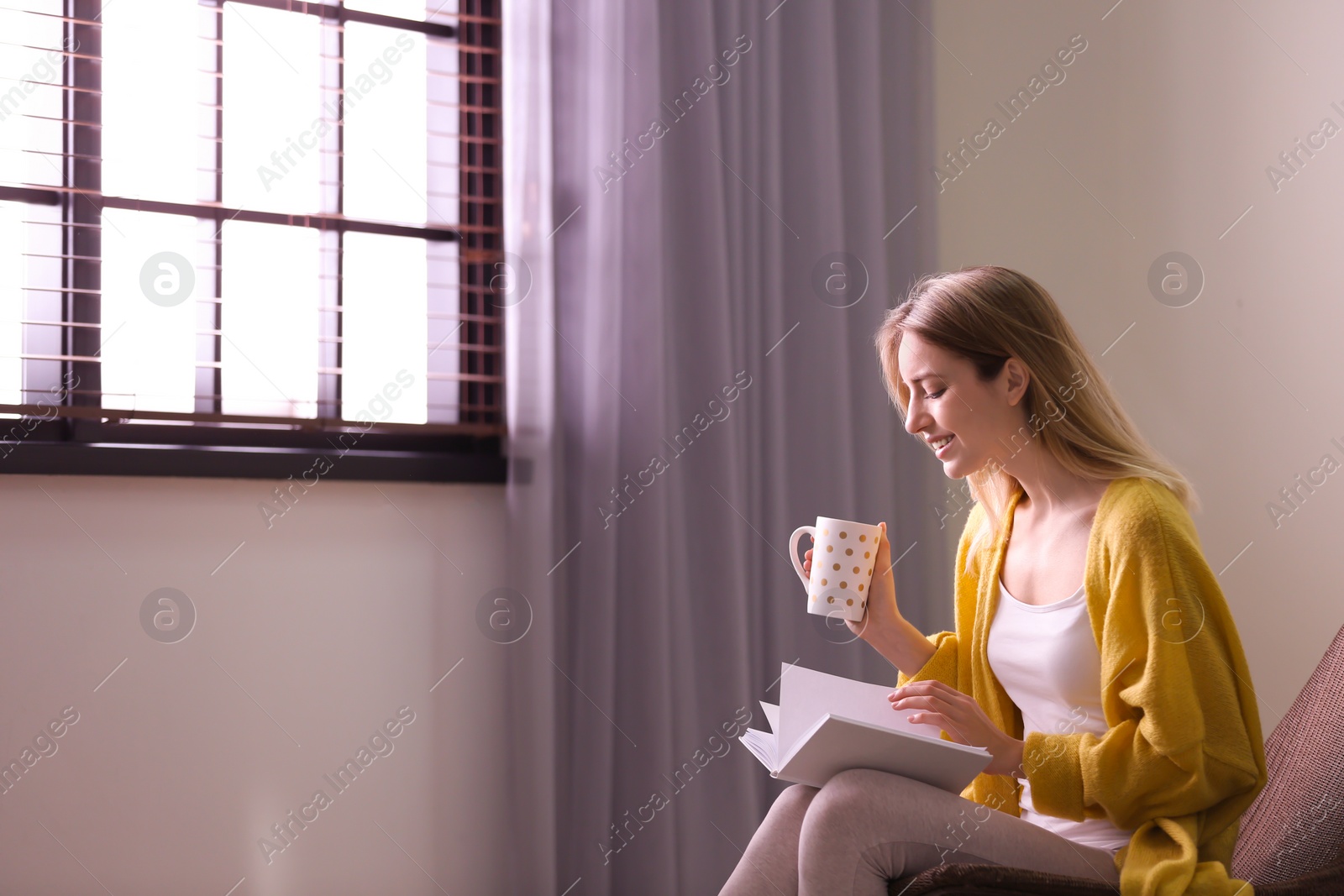 Photo of Young woman reading book near window with blinds at home. Space for text