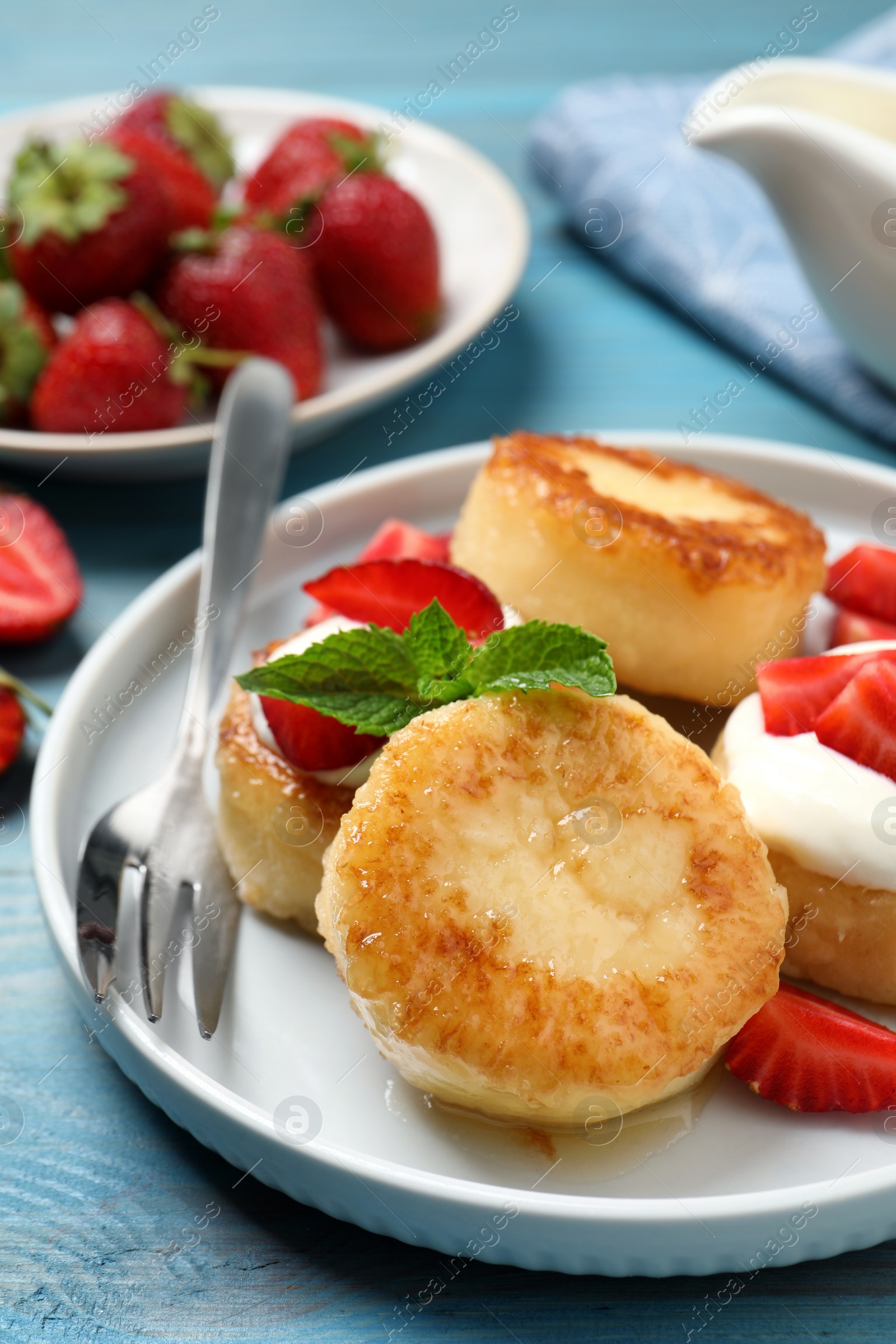 Photo of Delicious cottage cheese pancakes with strawberries, honey and sour cream on light blue wooden table, closeup