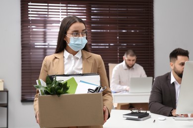 Upset dismissed woman wearing protective mask carrying box with personal stuff in office