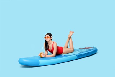 Happy woman with refreshing drink resting on SUP board against light blue background