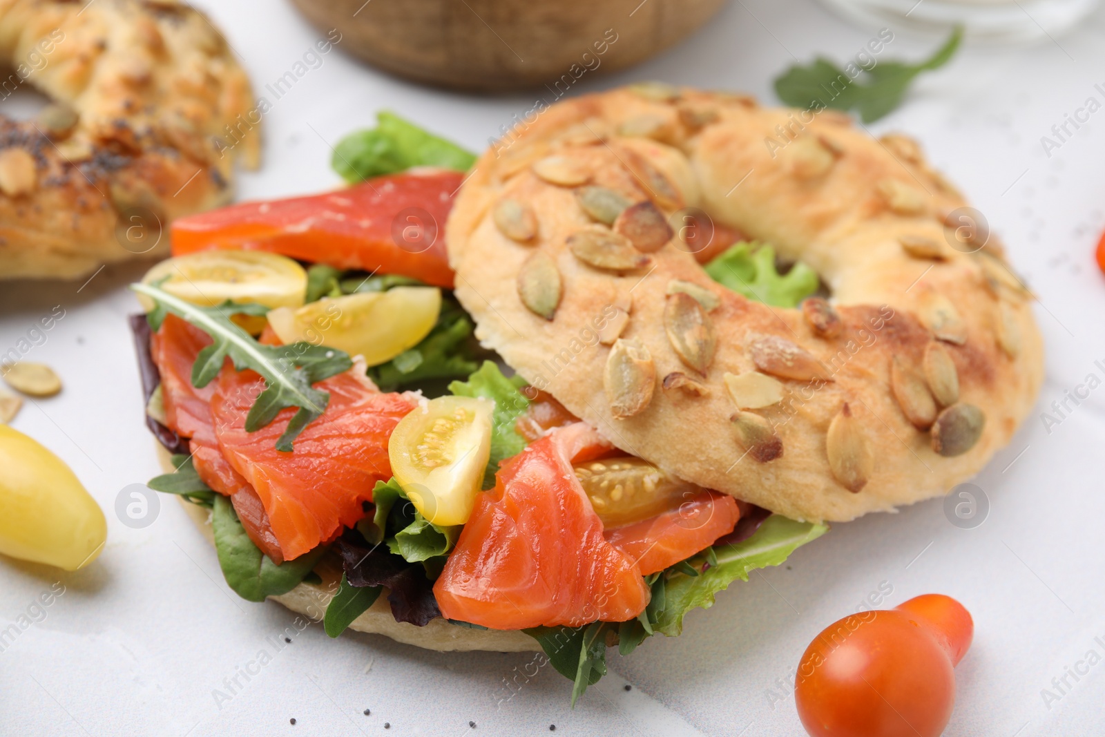 Photo of Tasty bagel with salmon, lettuce, tomatoes and arugula on white tiled table, closeup