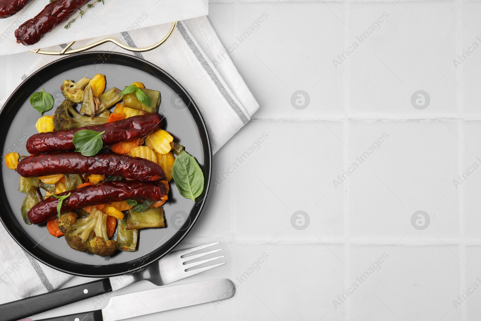 Photo of Delicious smoked sausages and baked vegetables served on white tiled table, flat lay. Space for text