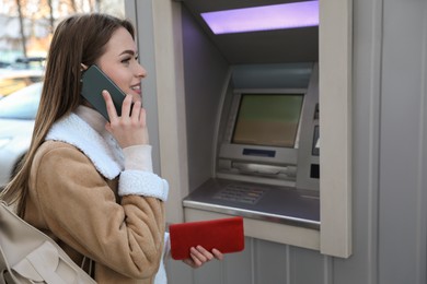 Photo of Young woman talking by mobile phone while using cash machine for money withdrawal outdoors