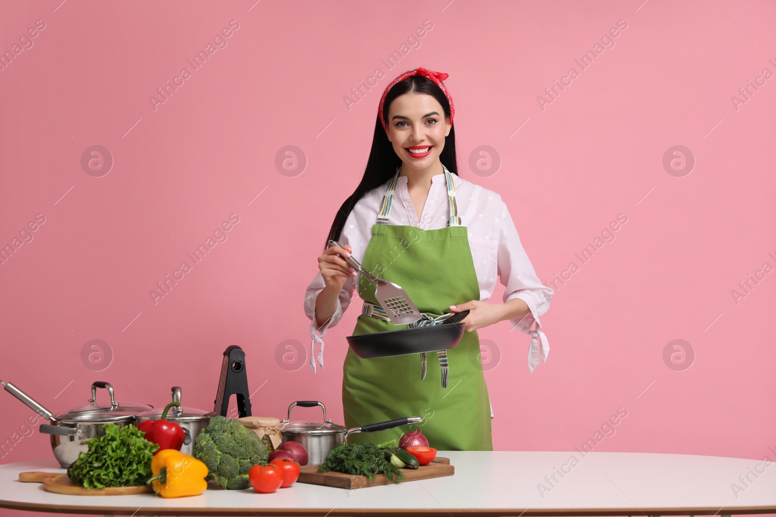 Photo of Young housewife with pan, spatula and products on pink background