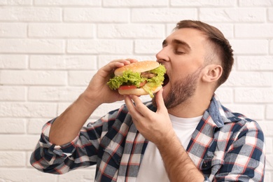 Young man eating tasty burger against brick wall