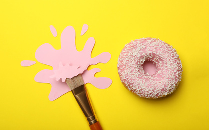Photo of Artist's brush with paint blot and donut on yellow background, flat lay