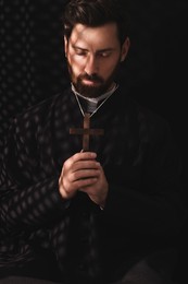 Photo of Catholic priest in cassock holding cross and praying to God in confessional