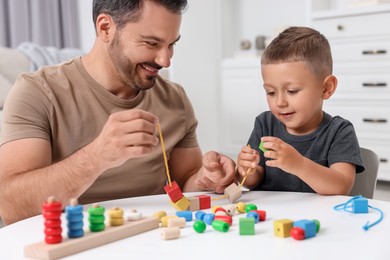 Motor skills development. Father and his son playing with wooden pieces and string for threading activity at table indoors