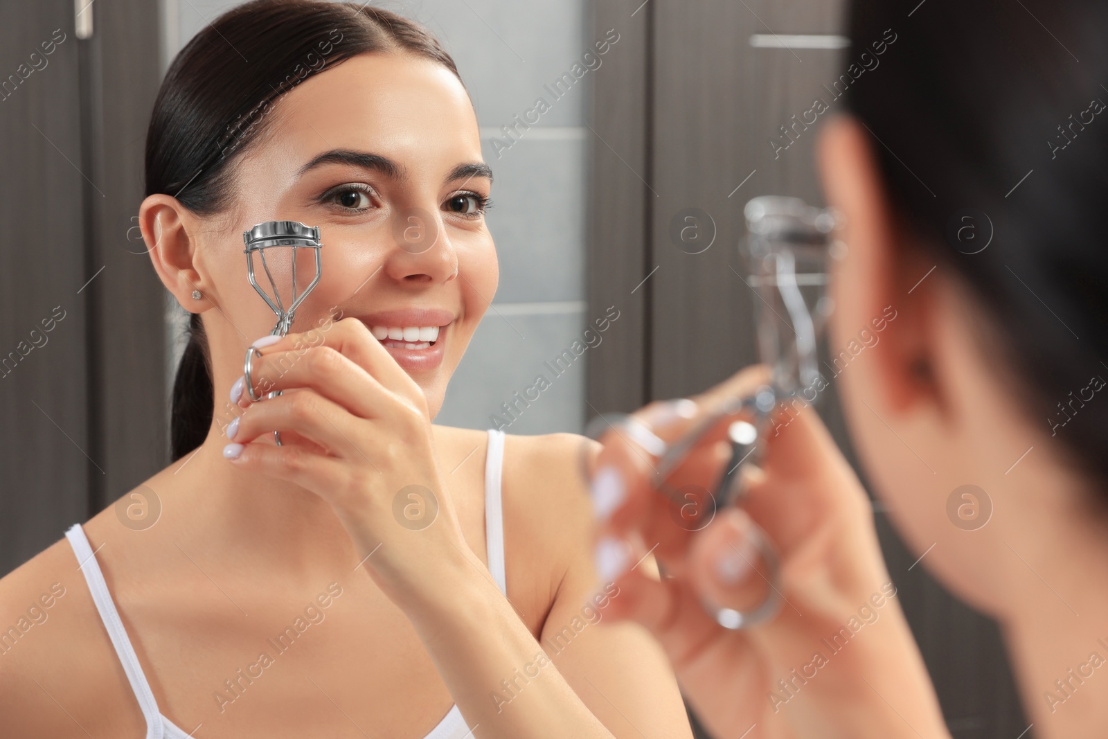 Photo of Beautiful young woman using eyelash curler near mirror in bathroom