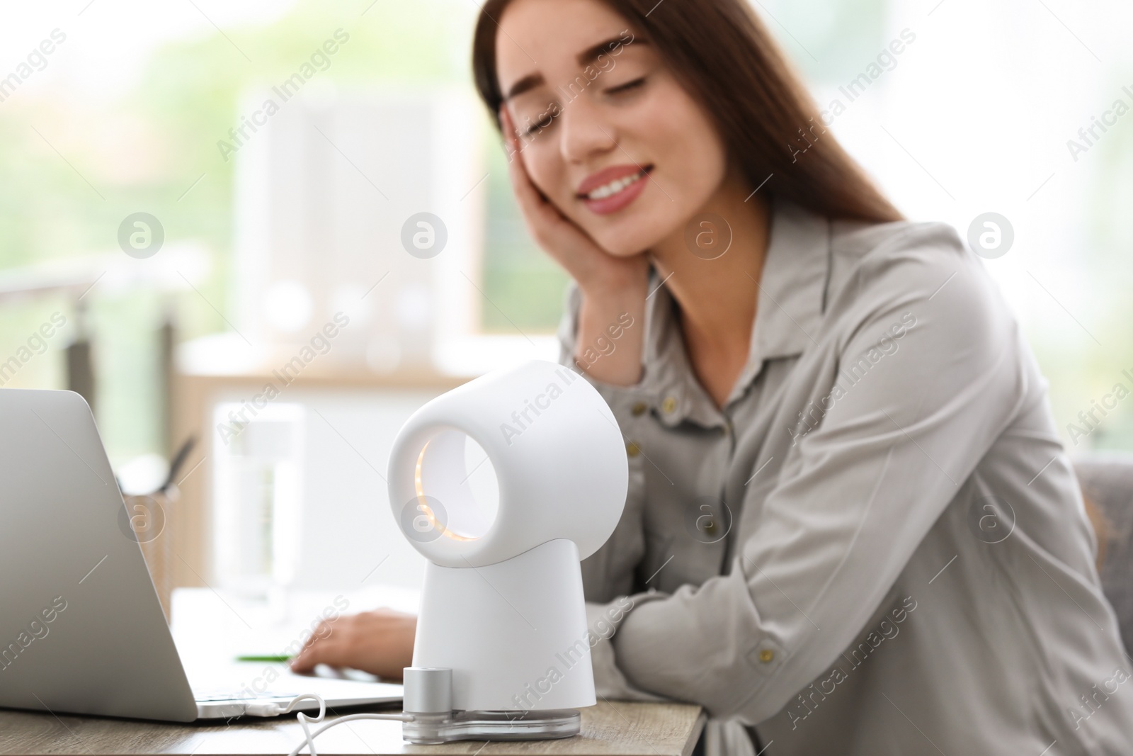 Photo of Young woman enjoying air flow from portable fan at workplace. Summer heat