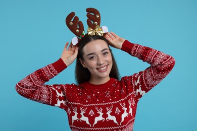 Photo of Young woman in Christmas sweater and reindeer headband on light blue background