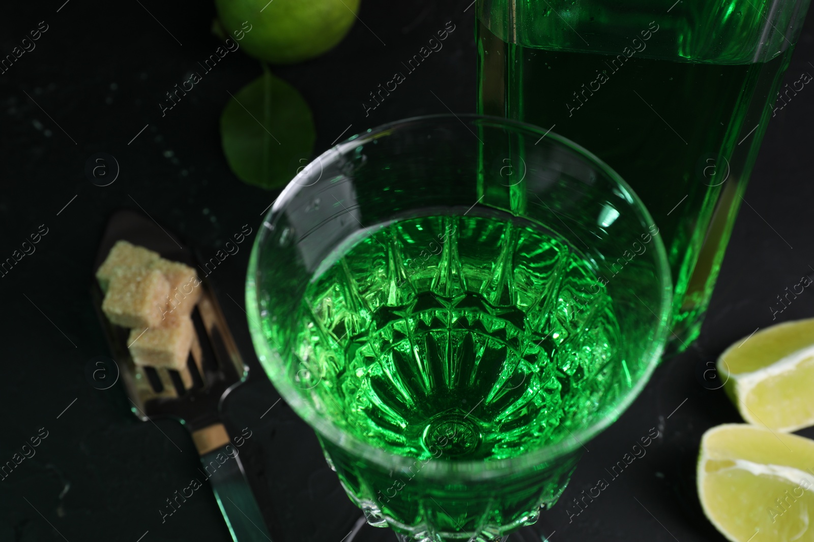 Photo of Absinthe in glass, spoon, brown sugar and lime on black table, closeup. Alcoholic drink