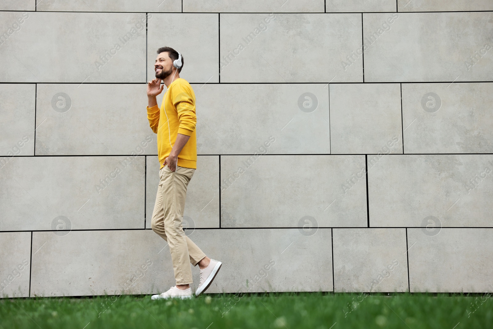 Photo of Handsome man with headphones walking near grey stone wall outdoors, space for text