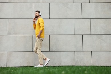 Handsome man with headphones walking near grey stone wall outdoors, space for text