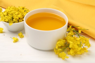 Photo of Rapeseed oil in bowl and beautiful yellow flowers on white wooden table