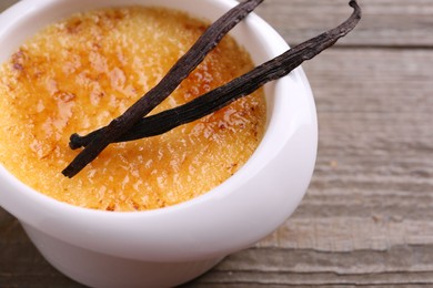Photo of Delicious creme brulee in bowl and vanilla pods on wooden table, closeup
