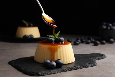 Photo of Pouring caramel onto delicious pudding with blueberries and mint on grey table against black background