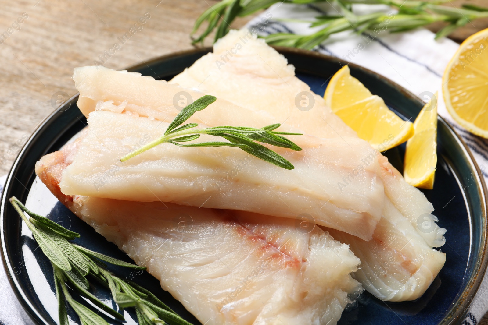 Photo of Pieces of raw cod fish, rosemary and lemon on wooden table, closeup