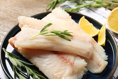 Photo of Pieces of raw cod fish, rosemary and lemon on wooden table, closeup