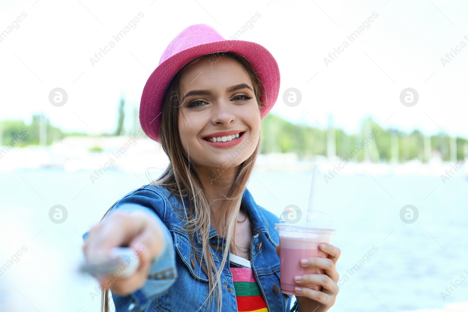 Photo of Happy young woman with drink taking selfie on riverside