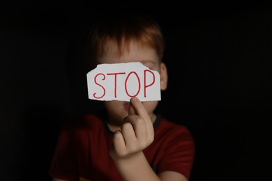 Photo of Little boy holding piecepaper with word Stop in darkness, focus on hand. Domestic violence concept